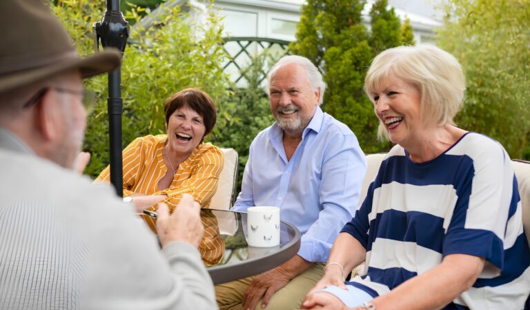 Four seniors smiling and chatting around a glass patio table in a sunny garden.