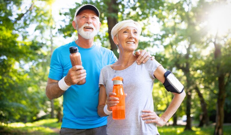 Smiling senior couple in a sunlit park, each holding a water bottle; man has his arm around the woman.