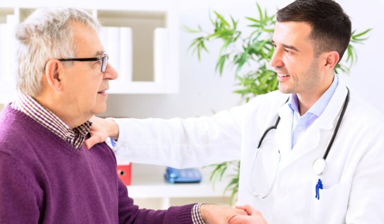 Doctor in a white coat and stethoscope speaks with an elderly man in a purple sweater in a bright clinic, clasping his hand.