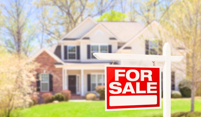 “For sale” sign in the foreground with a white post and red sign, a house and yard blurred in the background.